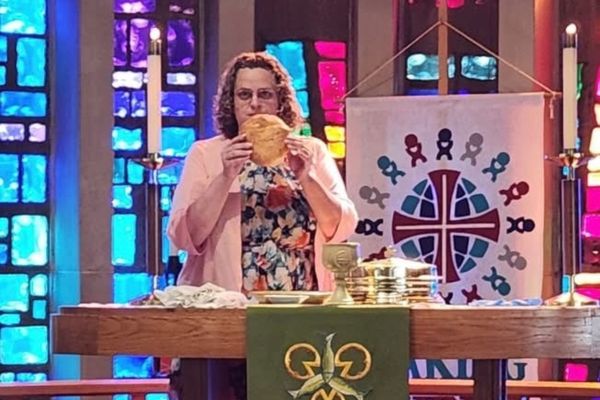 A person in a pink cardigan and floral dress stands at an altar, holding a piece of bread. Behind them is a stained-glass window and a banner with a cross and figures. Chalices and bread are on the altar.