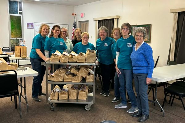 A group of people in teal shirts stand behind a rolling cart filled with brown paper lunch bags.