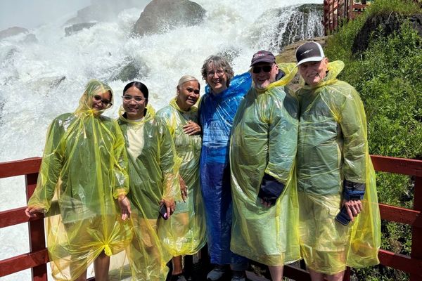 A group of six people wearing yellow ponchos stand on a wooden platform in front of a large waterfall.
