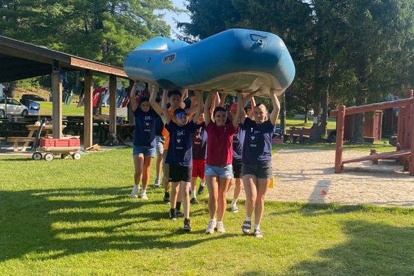 A group of people carrying a large blue inflatable raft over their heads on a grassy area with trees and a playground in the background.