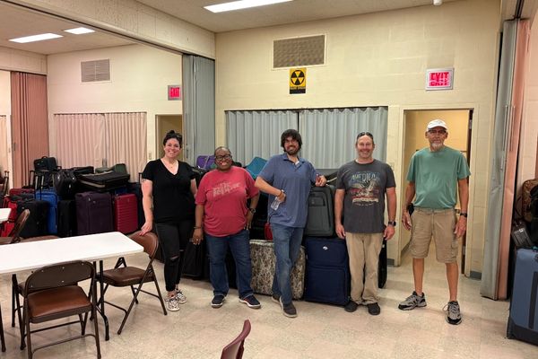 Five people stand in a room filled with luggage. Some of the luggage is stacked high behind them. A "radiation hazard" symbol is visible on the wall.
