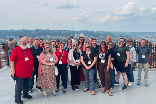 A group of people stand on a rooftop with a city skyline in the background under a cloudy sky.