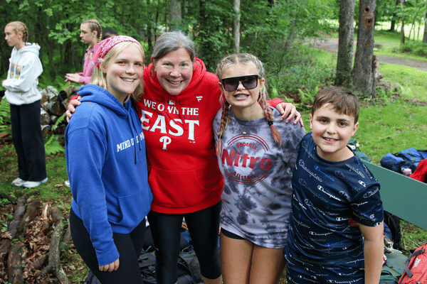 Four people stand outdoors on a path in a green park, smiling at the camera; one wears a red hoodie with “WE ON THE EAST” text, another a blue headband, and a child in a dark shirt stands at the front.