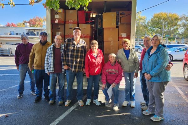 Group of people standing outdoors in front of a truck loaded with cardboard boxes and equipment, with trees and parked cars in the background.