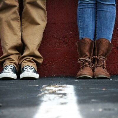 A close-up shot of two people's legs, one wearing tan pants and sneakers, the other wearing blue jeans and brown boots, standing against a red wall.